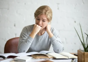 Kvinne som ser stresset ut med åpne bøker foran seg, et bilde som illustrerer forskjellen mellom akutt vs. kronisk stress i hverdagen.