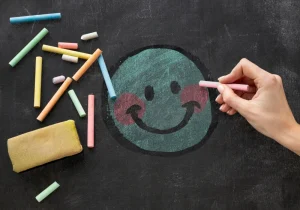 En hånd tegner et smilende ansikt med farget kritt på en tavle, omgitt av fargerike krittpinner – som symboliserer kreativitet og positiv angst gjennom uttrykk.