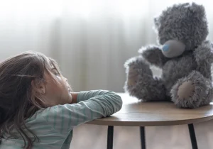 Et trist barn ser på en teddybjørn på den andre siden av et bord, noe som illustrerer angst hos barn og følelsen av ensomhet og usikkerhet i barndommen under stress.